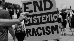 Black & white photo - hand holding carboard sign "END SYSTEMIC RACISM"
