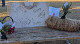 Objects on a wooden picnic table: small basket, hand-drawn sign reading "Get a kid's camp Yule Log", logs bundled in burlap with an evergreen sprig sticking up