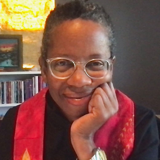 Rev. Patrice Curtis smiling at camera, chin on hand, wearing a dark robe and a red and gold stole