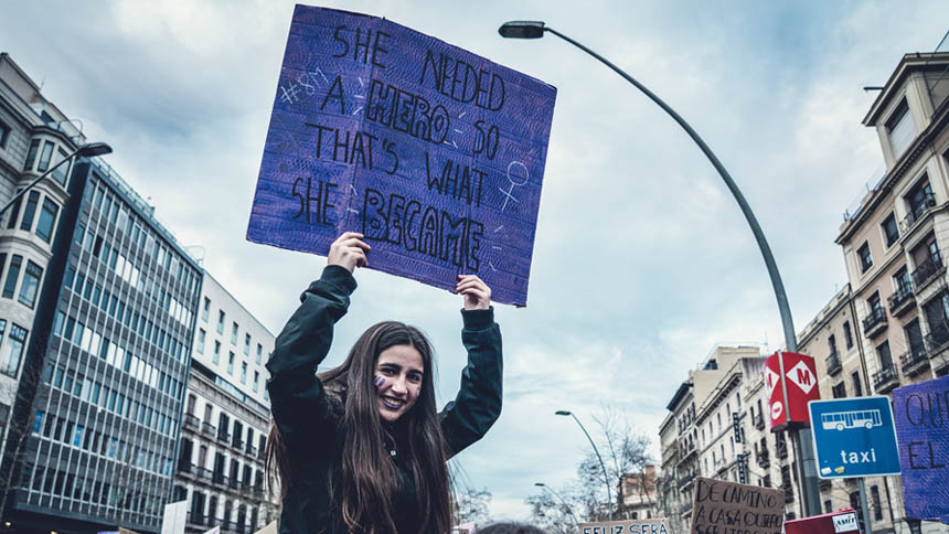 Young woman with long dark hair in a street protest, holding a sign "She needed a hero so that's what she became"